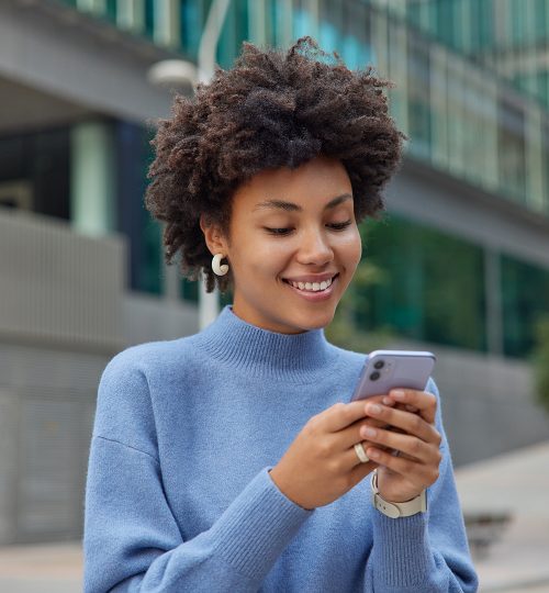 Glad young woman with curly bushy hair holds modern mobile phone downloads amazing application surfs social networks wears casual blue jumper smiles positively poses against blurred background